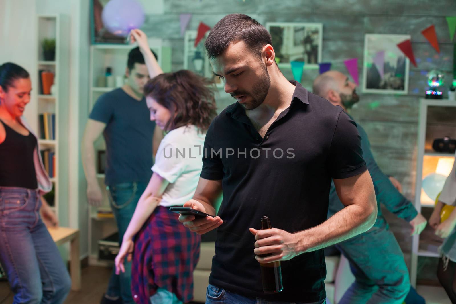 Young man using his smartphone at a party while all the beautiful females are dancing in the background.