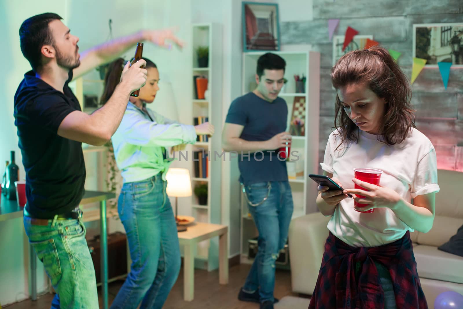 Cheerful young woman sitting in the centre of living room using her smartphone. Young people dancing.