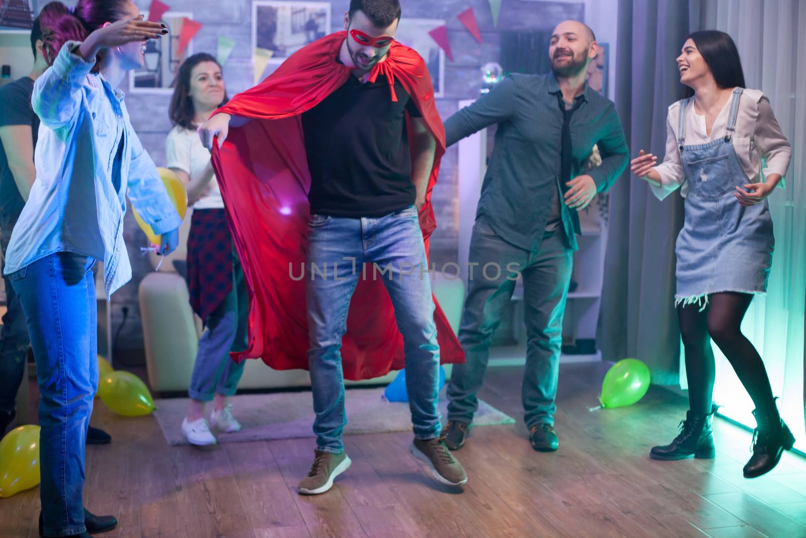 Young man wearing a superhero costume in a room with balloons dancing with his friends.