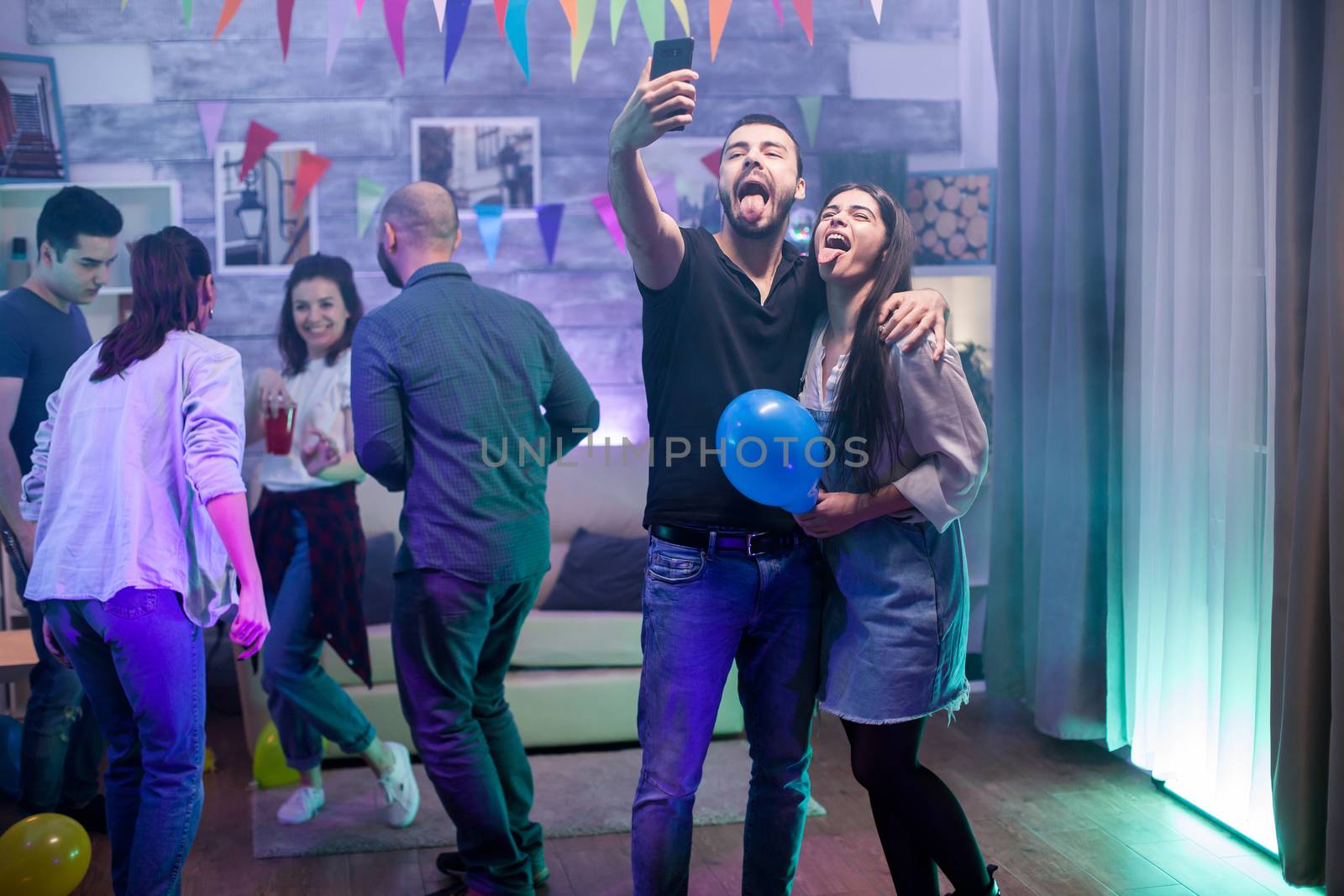 Bearded man and his girlfriend with tongue out taking a selfie while partying with their friends.