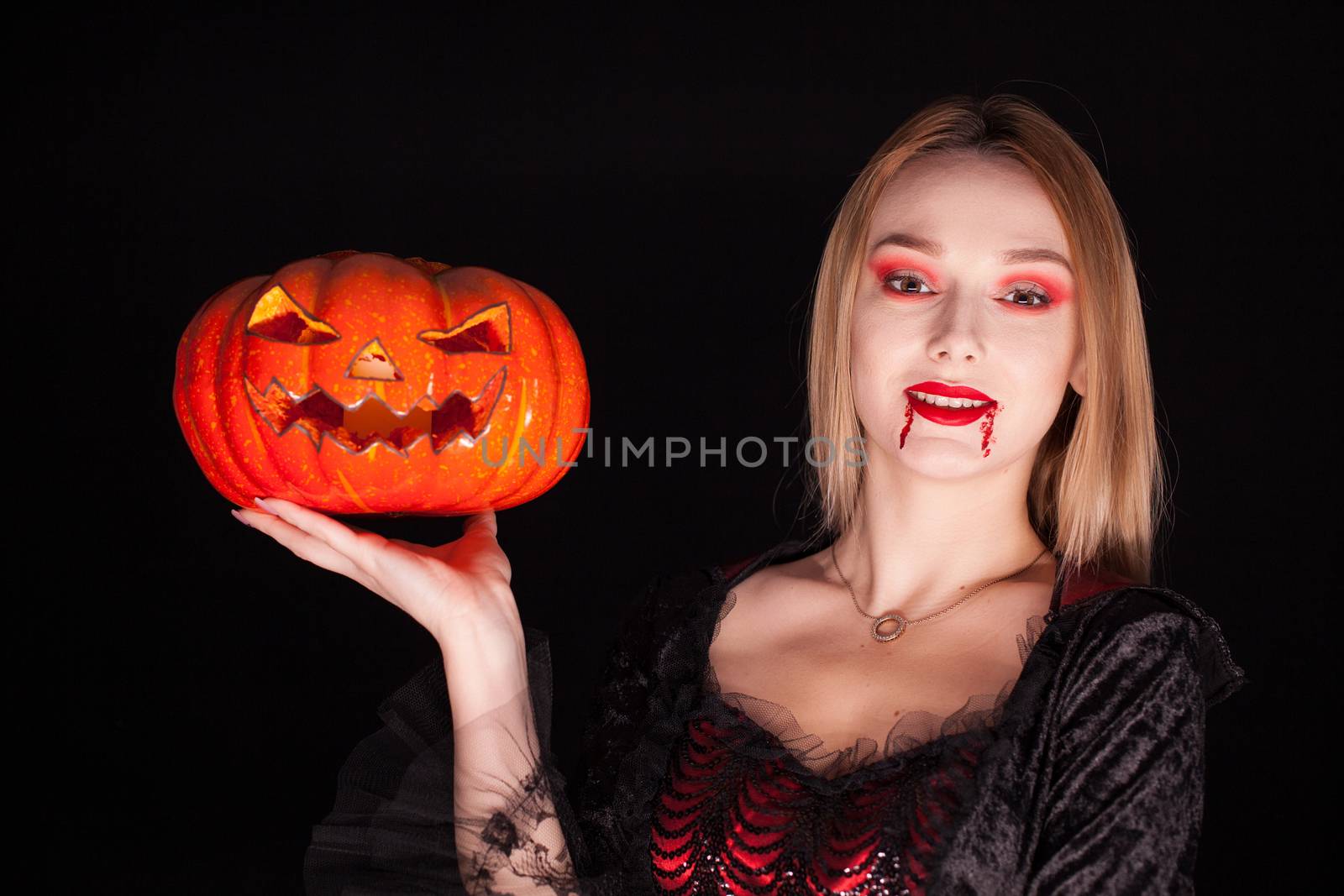 Portrait of beautiful woman dressed up like a vampire with bloody lips holding a pumpkin over black background.