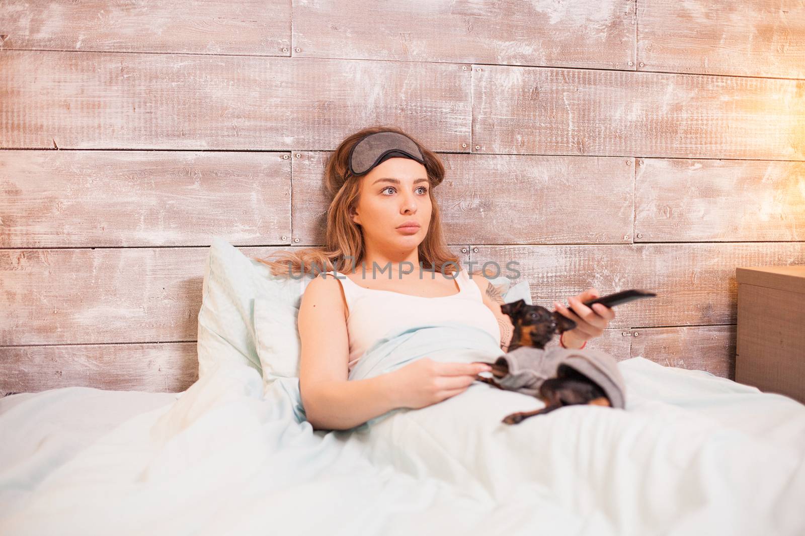 Beautiful young woman with eye cover mask watching tv in bed with her little dog.