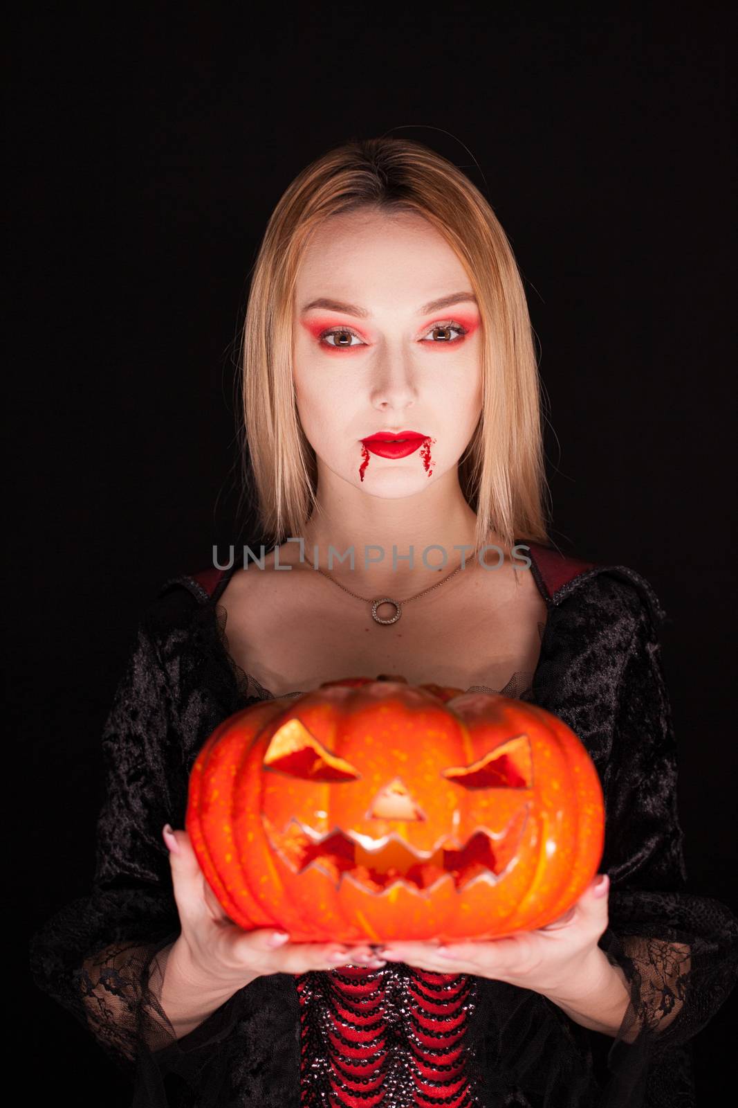 Beautiful girl dressed up like a vampire holding a pumpkin for halloween over black background.
