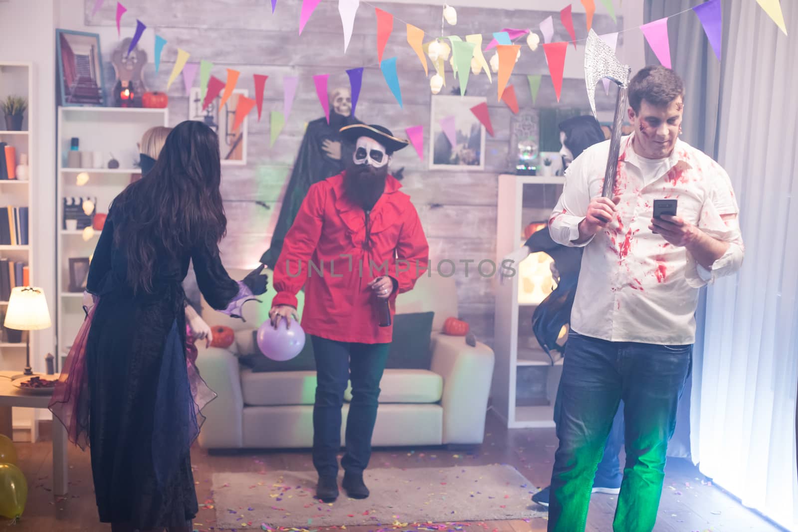 Young man dressed up like a zombie with an axe scrolling on his phone at halloween party.