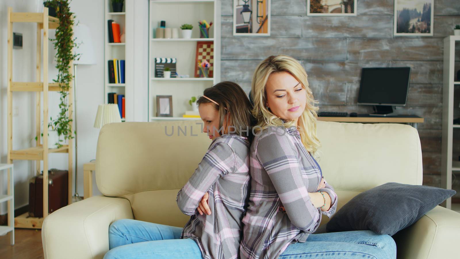 Little girl with bad behavior sitting back to back with her mother on the couch after a dispute.