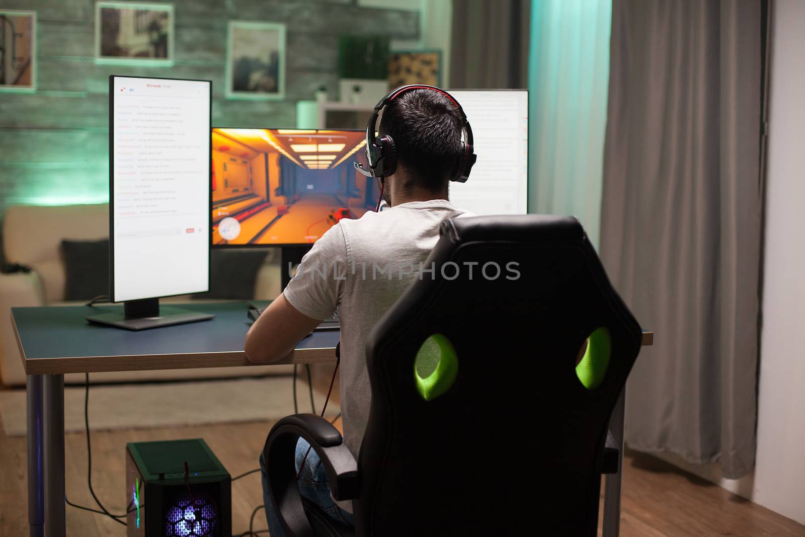 Back view of man sitting on gaming chair playing shooter game in the night. Neon light in the room.