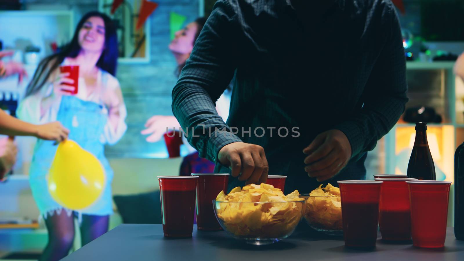 Young man taking chips from the table while his friends are dancing at the party in a room with neon lights and disco ball