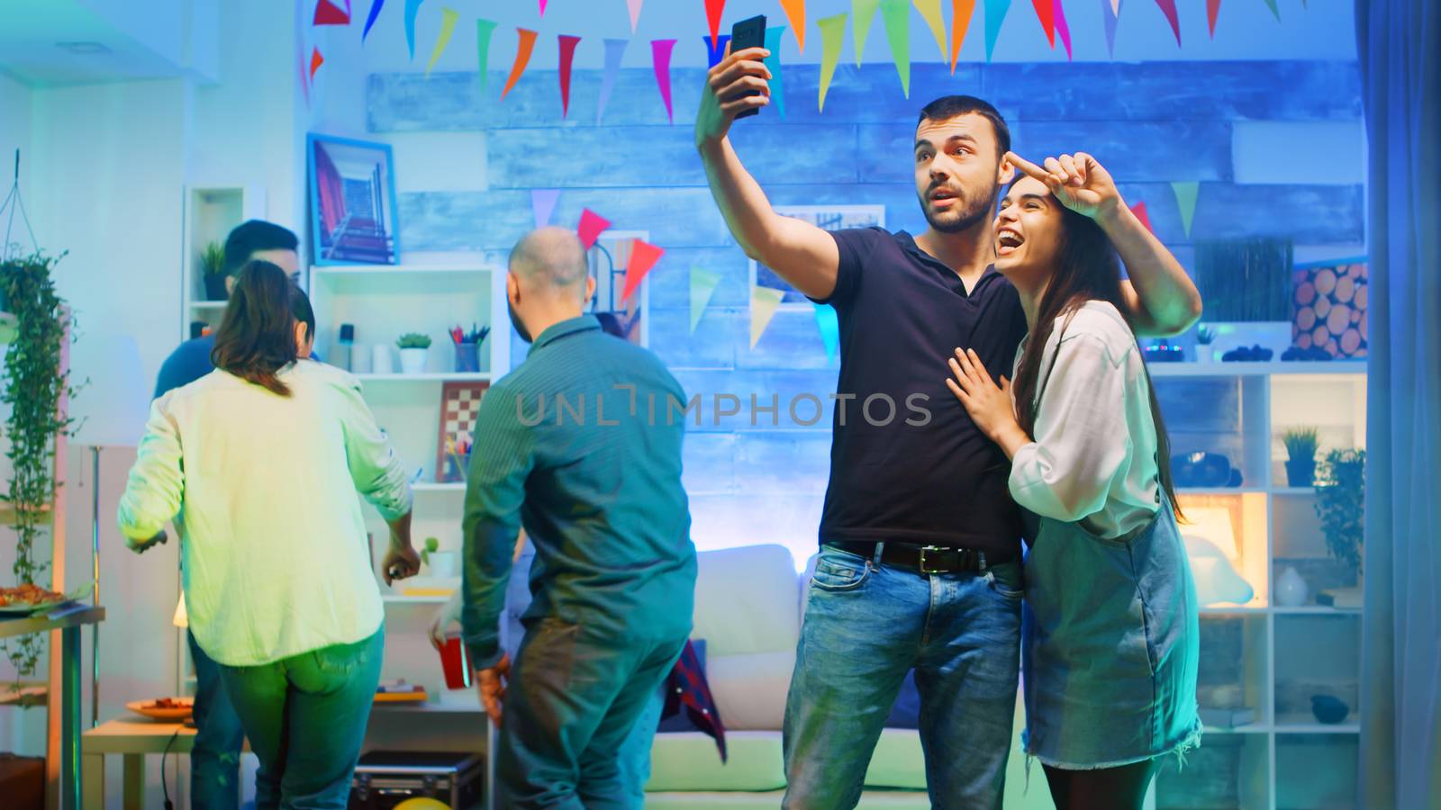 Attractive bearded young man taking a selfie with beautiful friend at the party. Loud music at wild college party in a room with neon lights, disco ball and a lot of alcohol