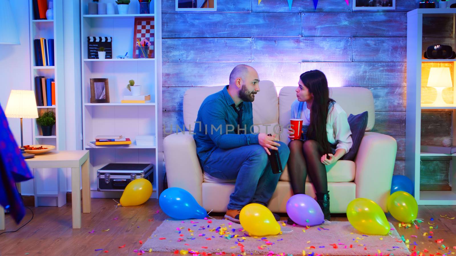 Group of people dancing at a party with neon lights in apartment while young couple is having an argument.