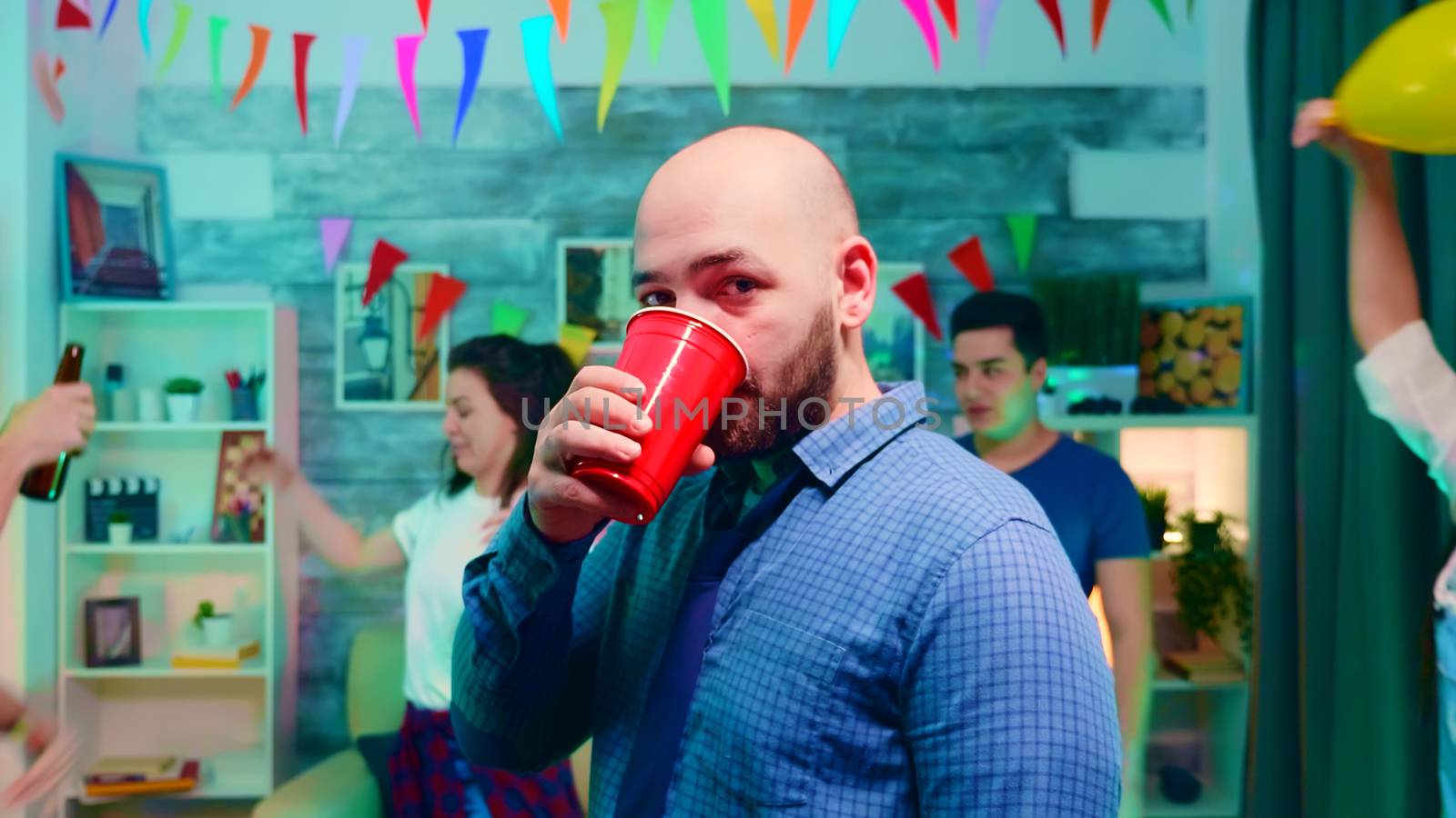 Zoom in shot of young man drinking alcohol while partying with his friends at college party with neon lights