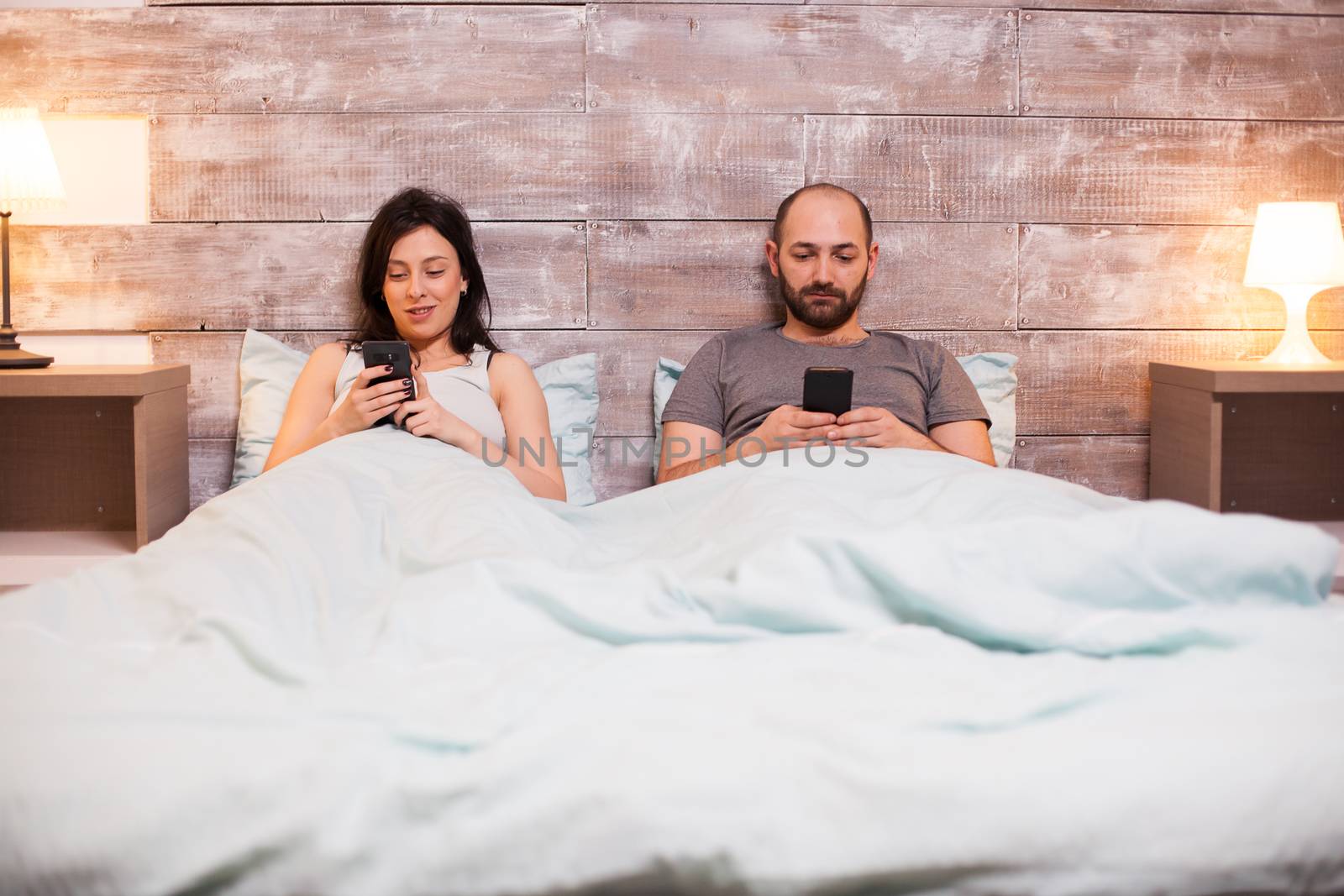 Caucasian couple in pajamas relaxing using smartphone before bedtime.
