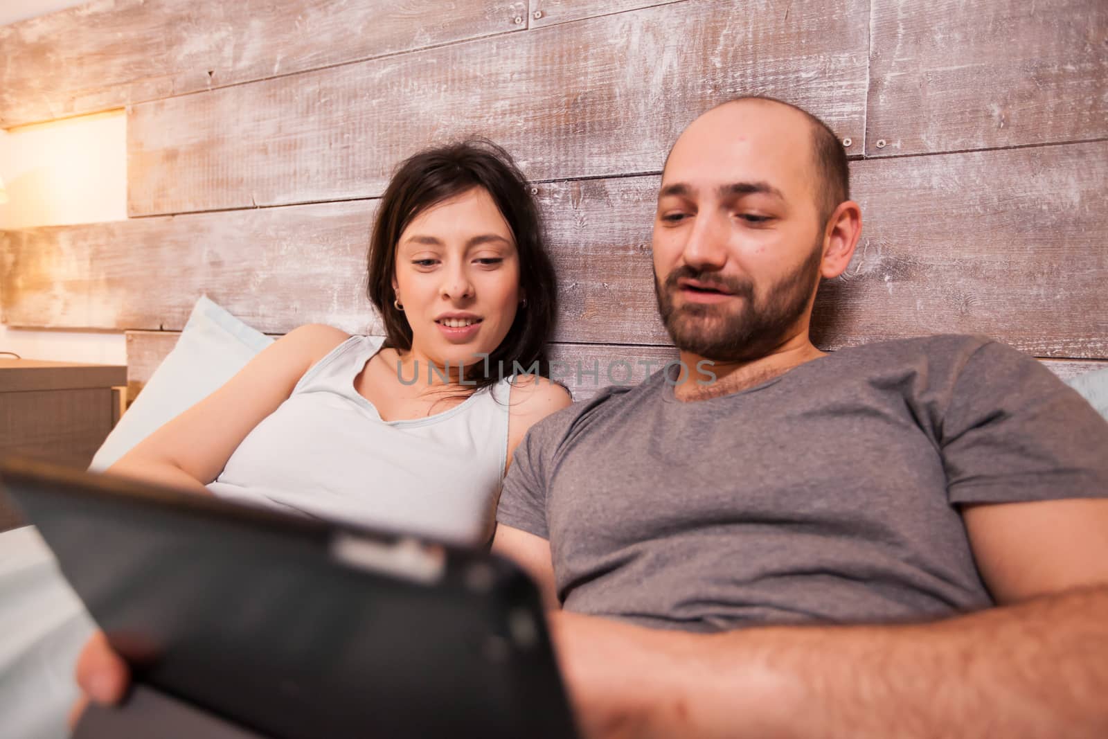 Happy boyfriend and girlfriend wearing pajamas lying in bed browsing on tablet computer.