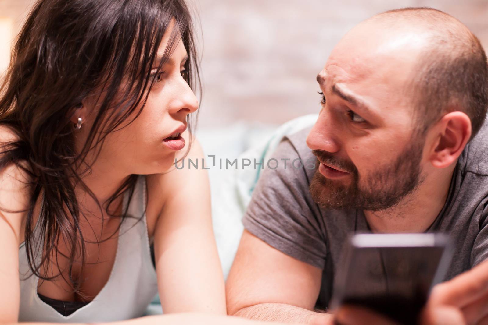 Boyfriend in pajamas looking worried at his girlfriend while using smartphone.