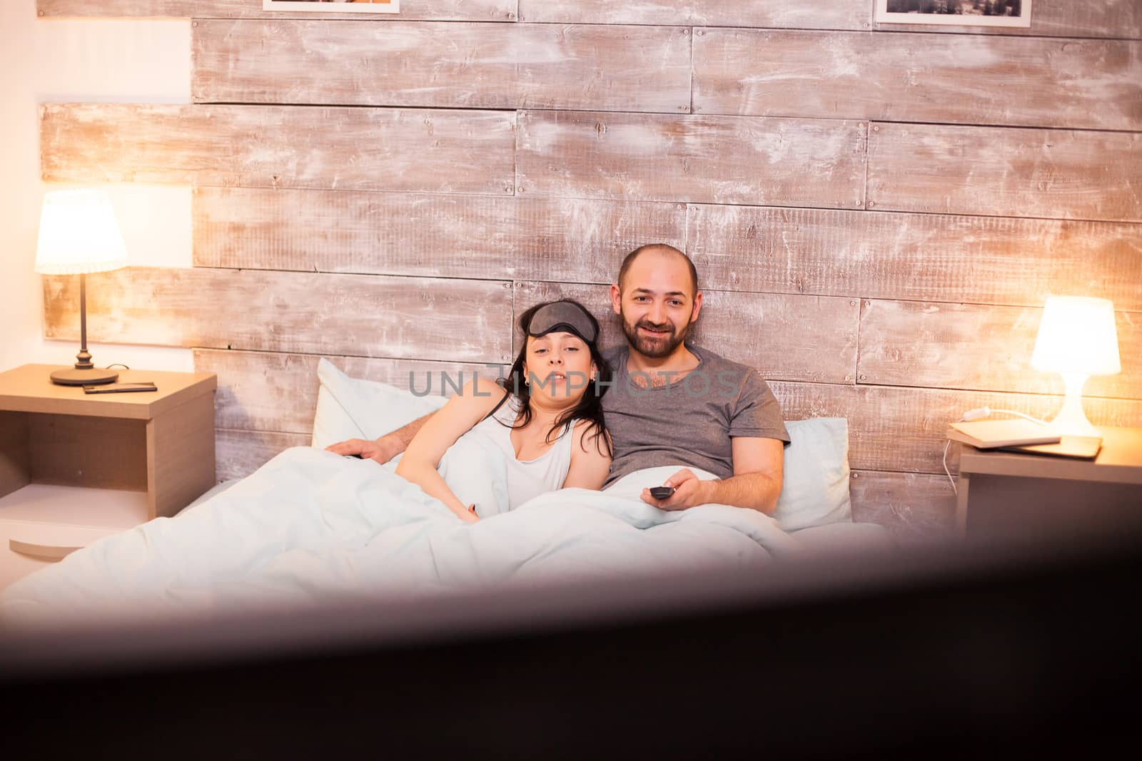 Young couple in pajamas watching tv lying under the blanket on comfortable bed.