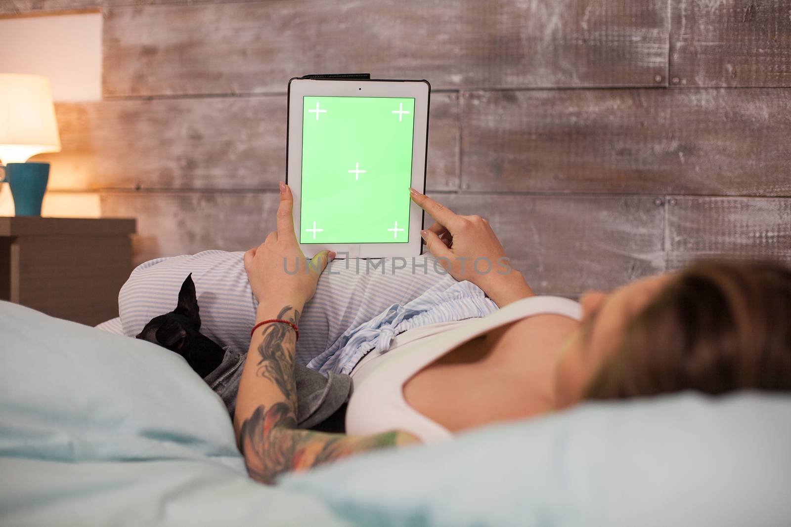Young woman lying in bed wearing pajamas pointing at tablet computer with green screen.