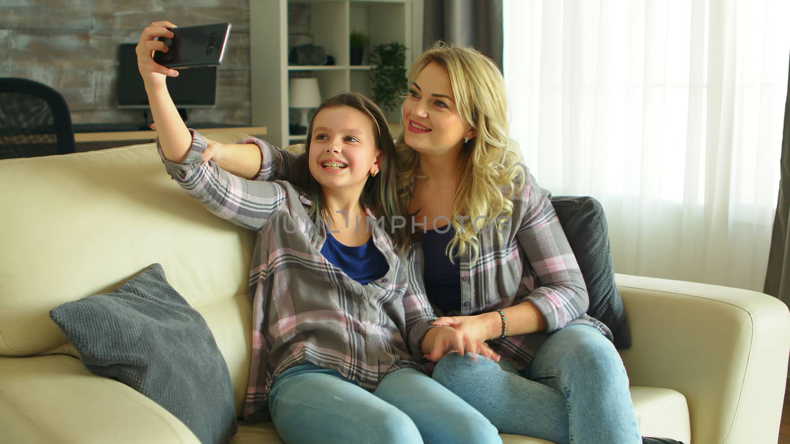 Mother and daughter making funny faces while taking a selfie sitting on the couch in living room.