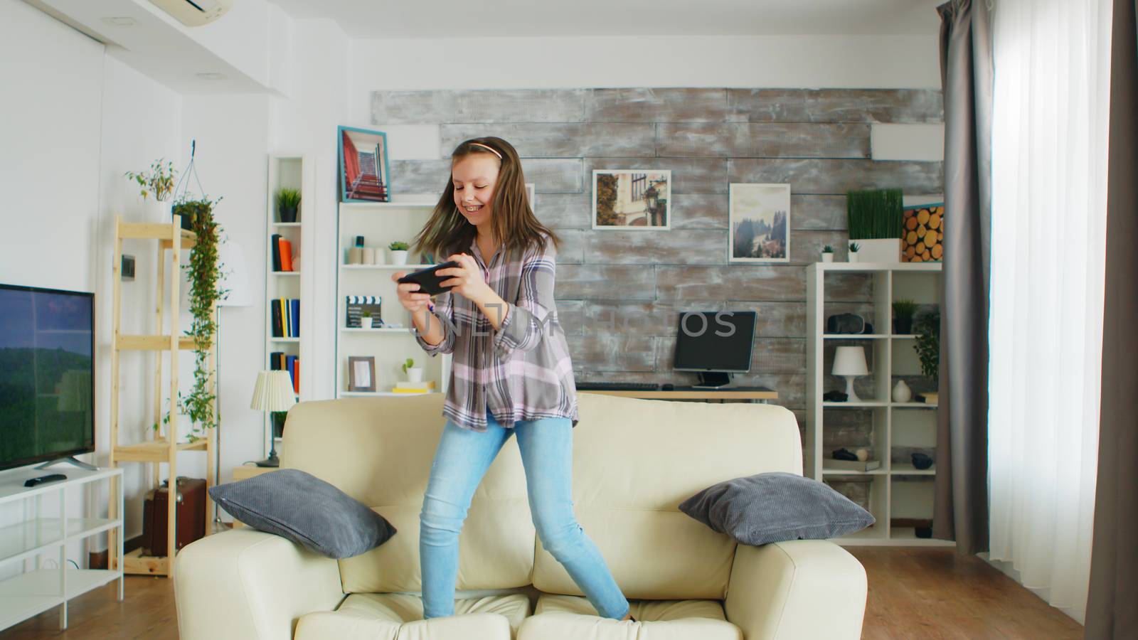 Little girl jumping on the couch in living room while playing video games on her phone.