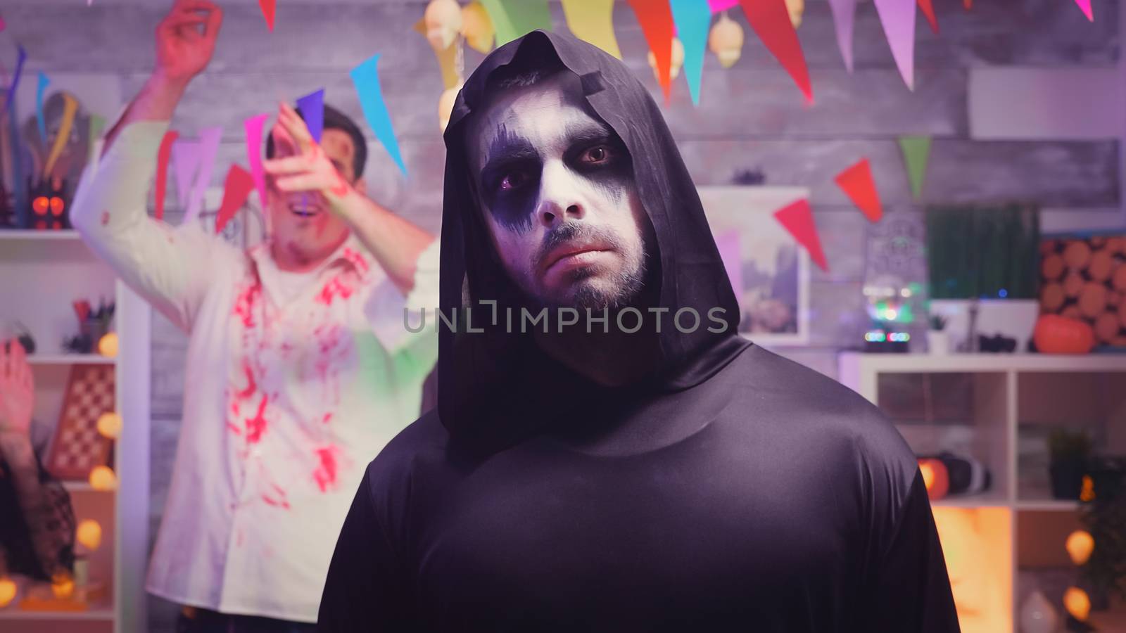 Zoom in shot of grim reaper at halloween party with his spooky friends dancing and having fun in the background