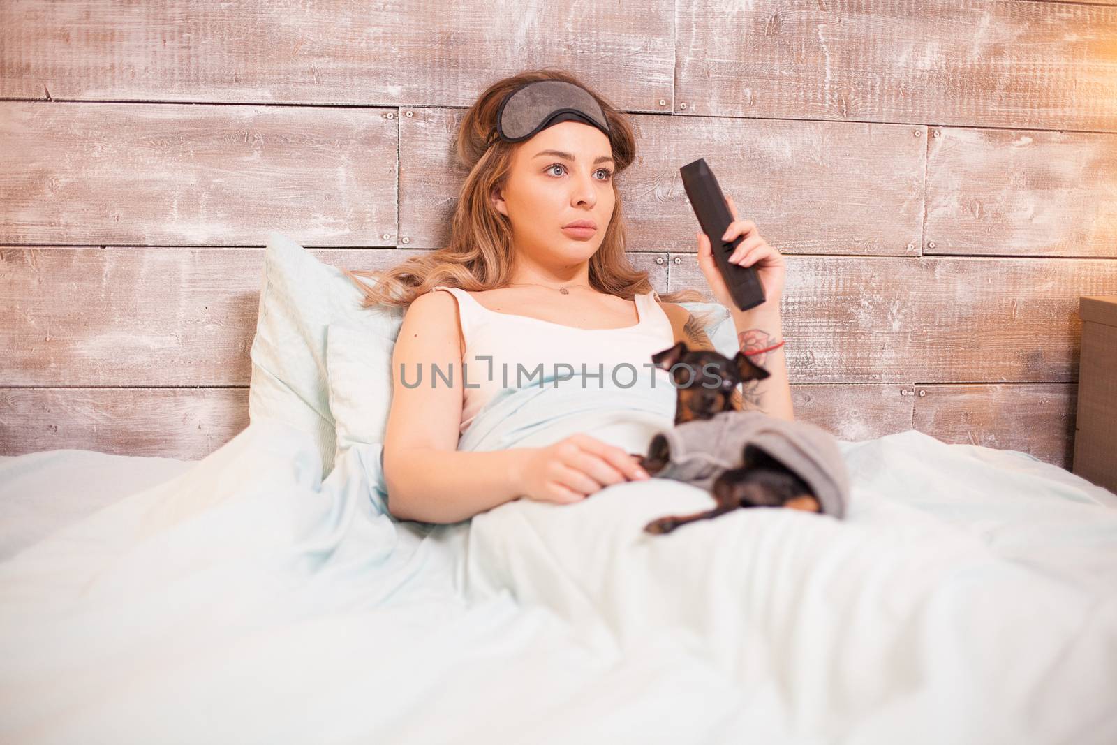 Calm beautiful woman with eye cover mask holding tv remote control with little dog.