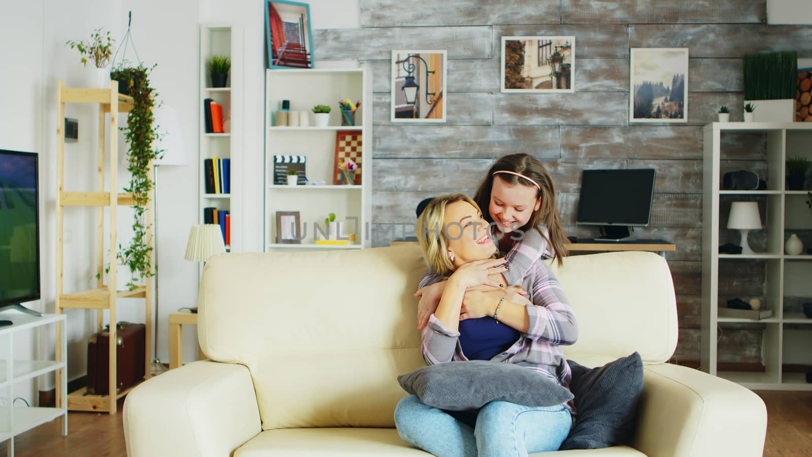 Happy little girl hugging her mother from behind while she is sitting on the couch in living room.