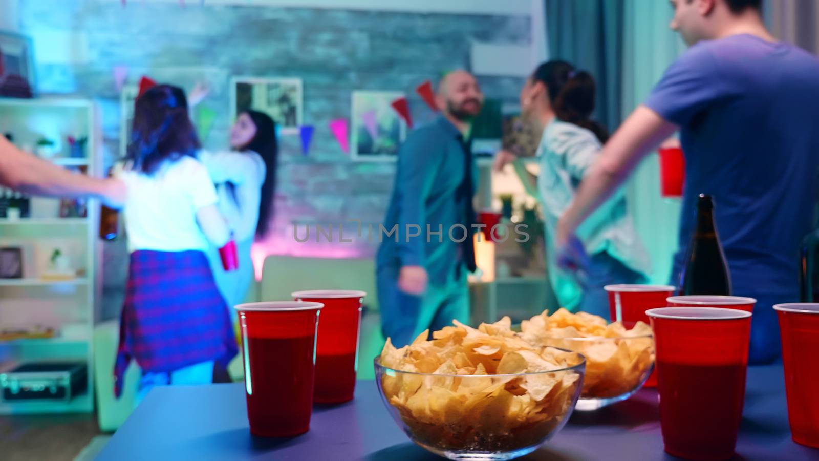 Young woman at a party with neon light taking chips from the table and a cup of beer by DCStudio