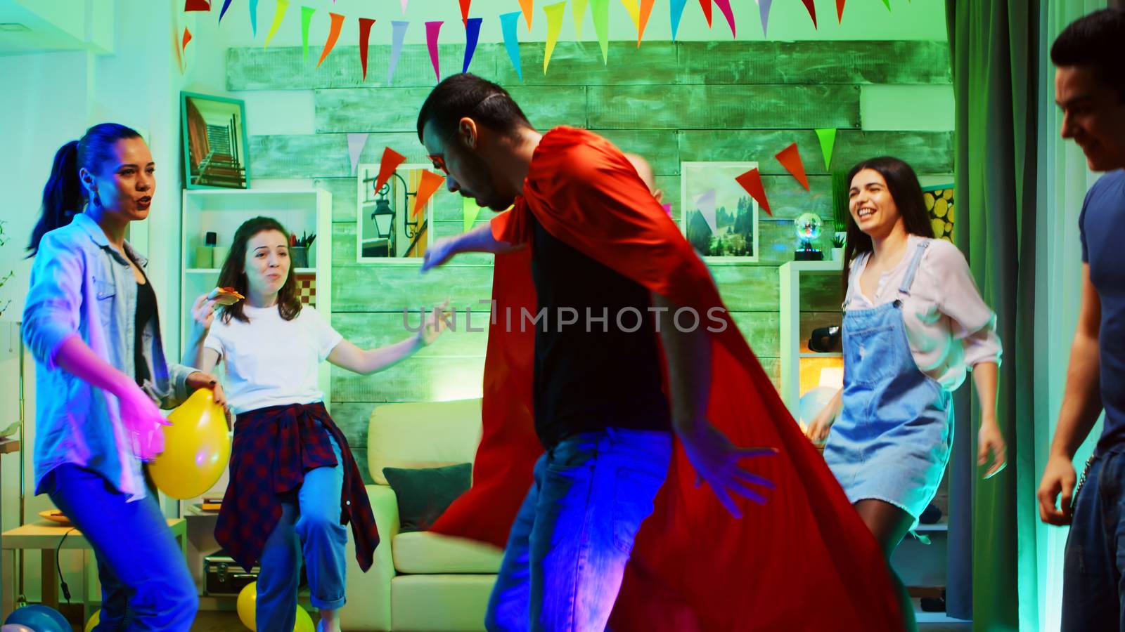 Handsome young man arriving at the party wearing a superhero red cape. Wild college party with neon lights, disco ball and alcohol