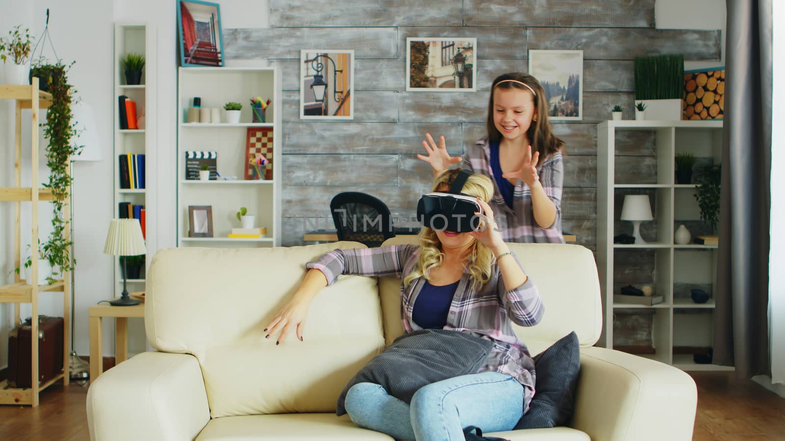 Little girl jumping around her mother while she's using virtual reality headset sitting on the couch.