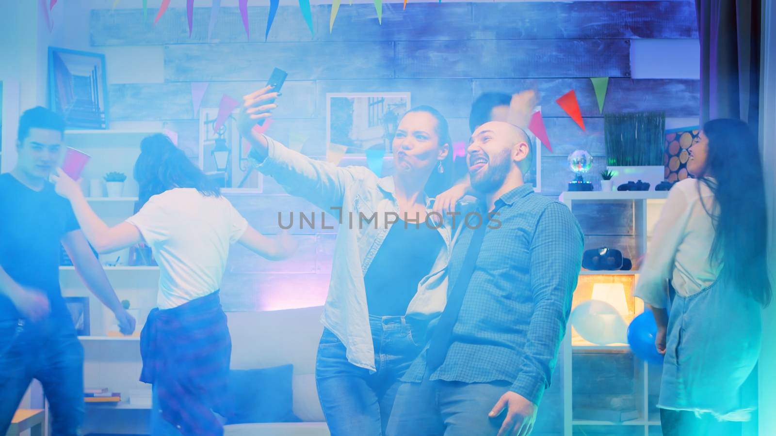 Young man and woman making funny faces while taking selfies at a party with neon lights and smoke.