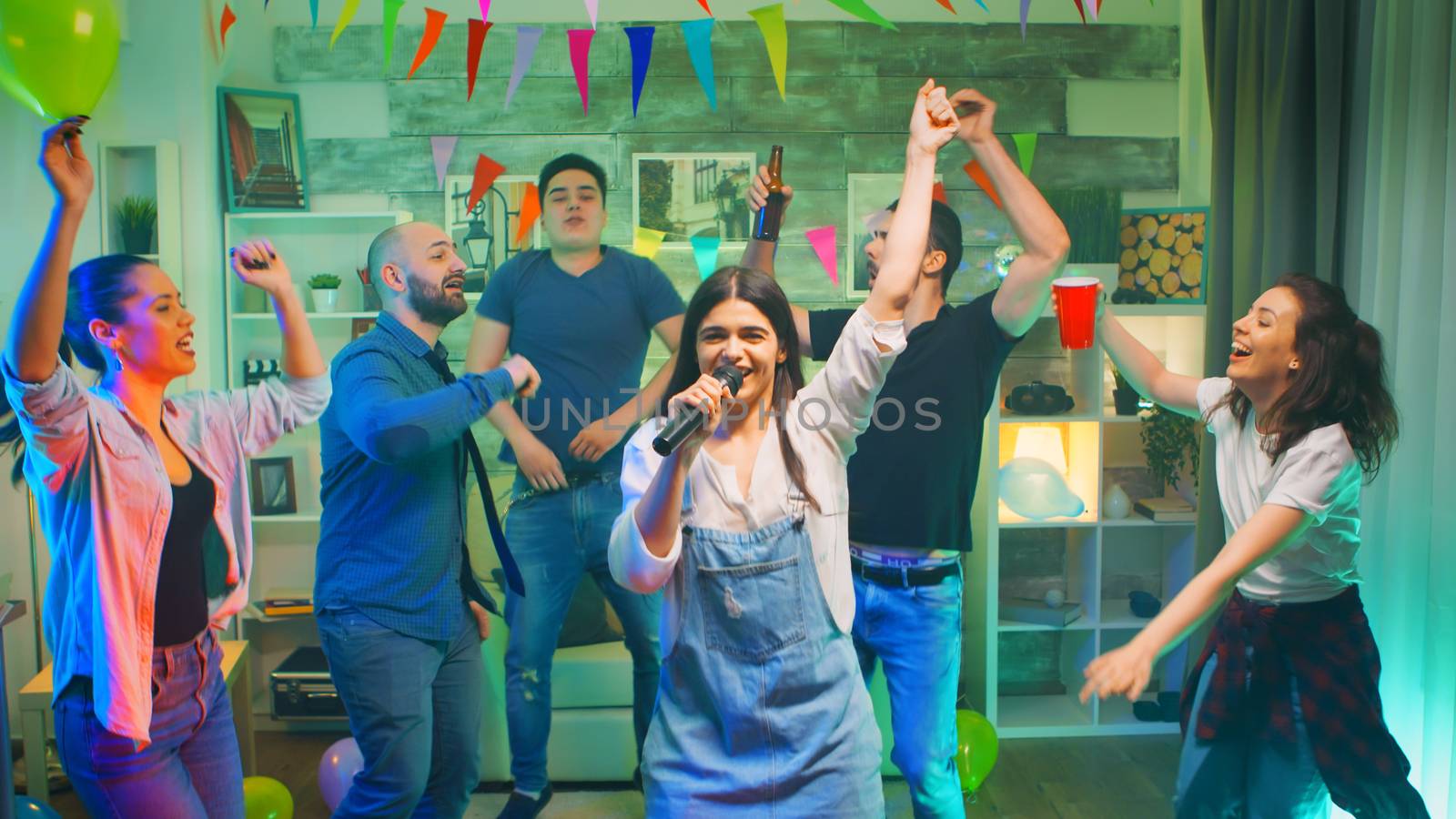 Beautiful young woman singing a song on microphone while partying with her group of friends in a room with neon lights, disco ball and alcohol