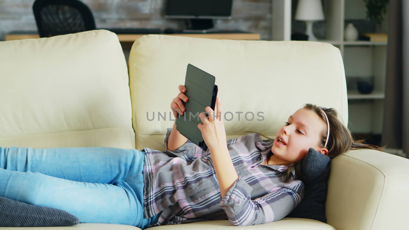 Little girl lying on the couch using her tablet. Cheerful kid.