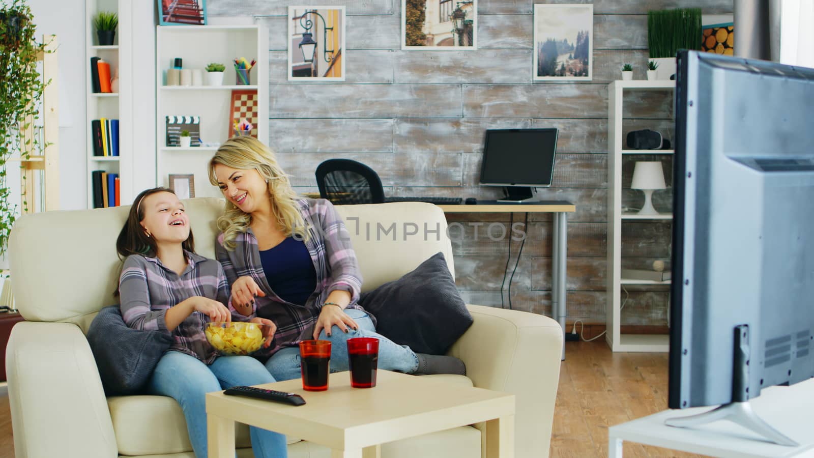 Little girl eating chips while watching tv with her mother sitting on the couch.