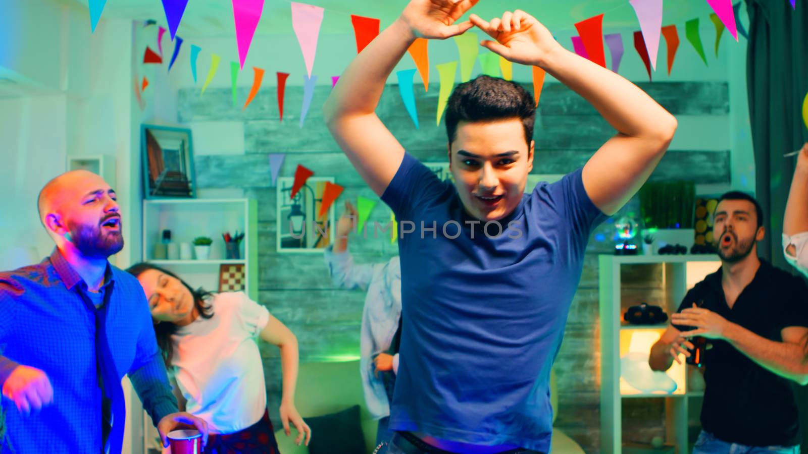 Cheerful young man dancing with his hands up at the party with his friends in an apartment with neon lights.