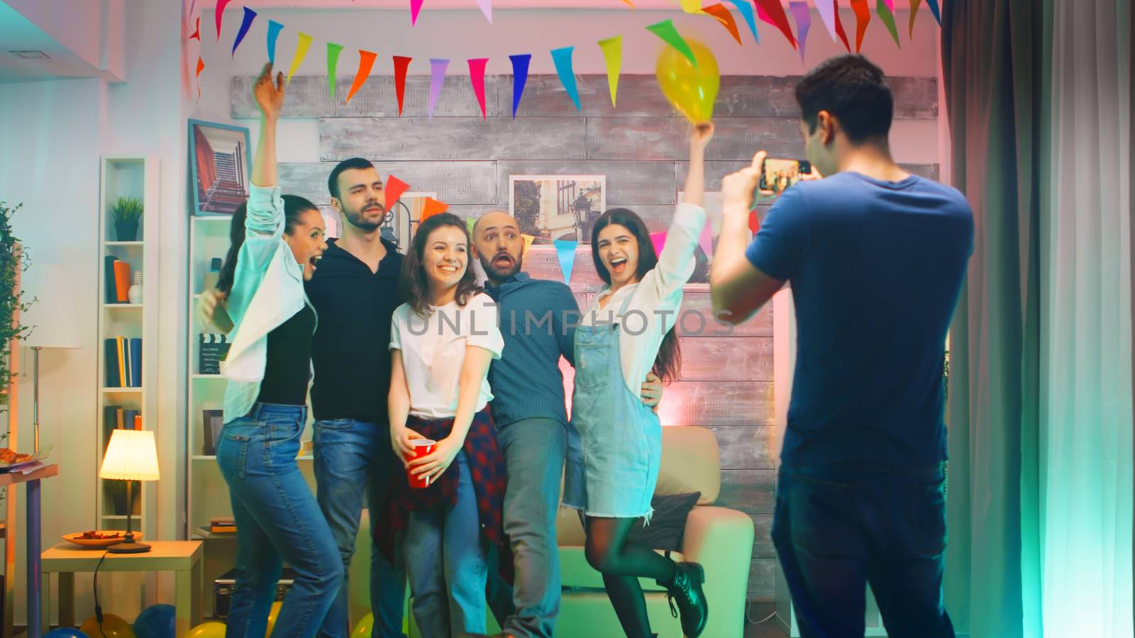 Beautiful young woman blowing kisses surrounded by friends while man taking group photos at the party with smartphone.