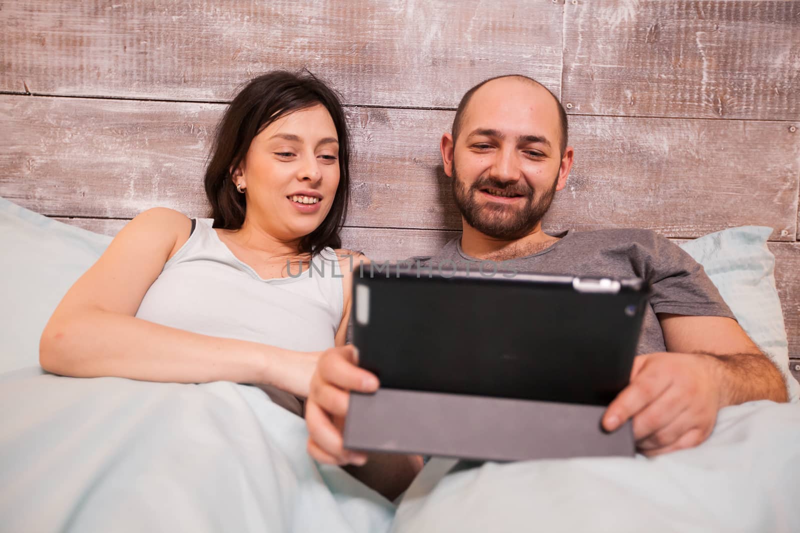 Beautiful young couple wearing pajamas sitting side by side in bed at night using tablet computer.