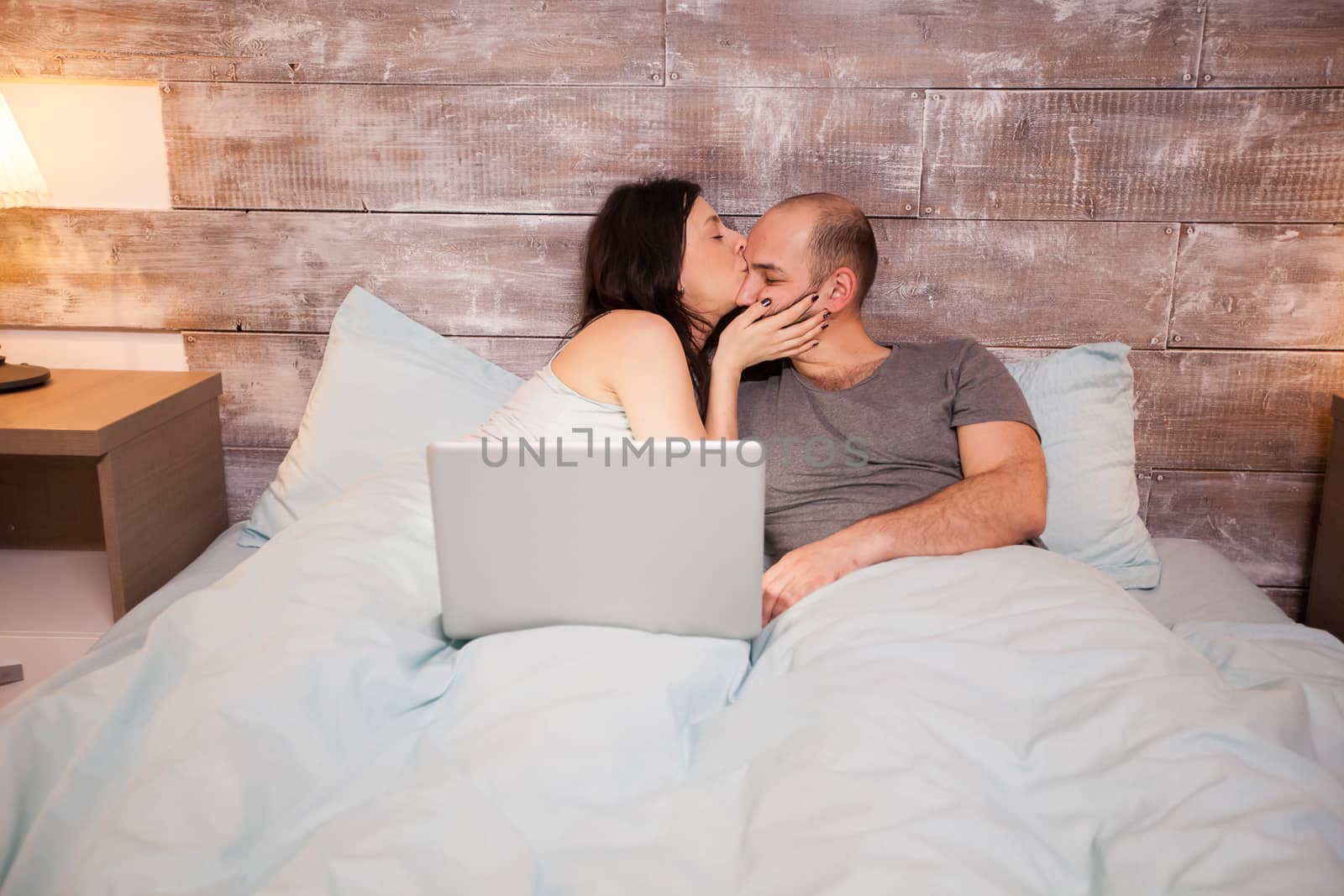Beautiful wife in pajamas kissing her beautiful husband before bedtime. Laptop on bed.