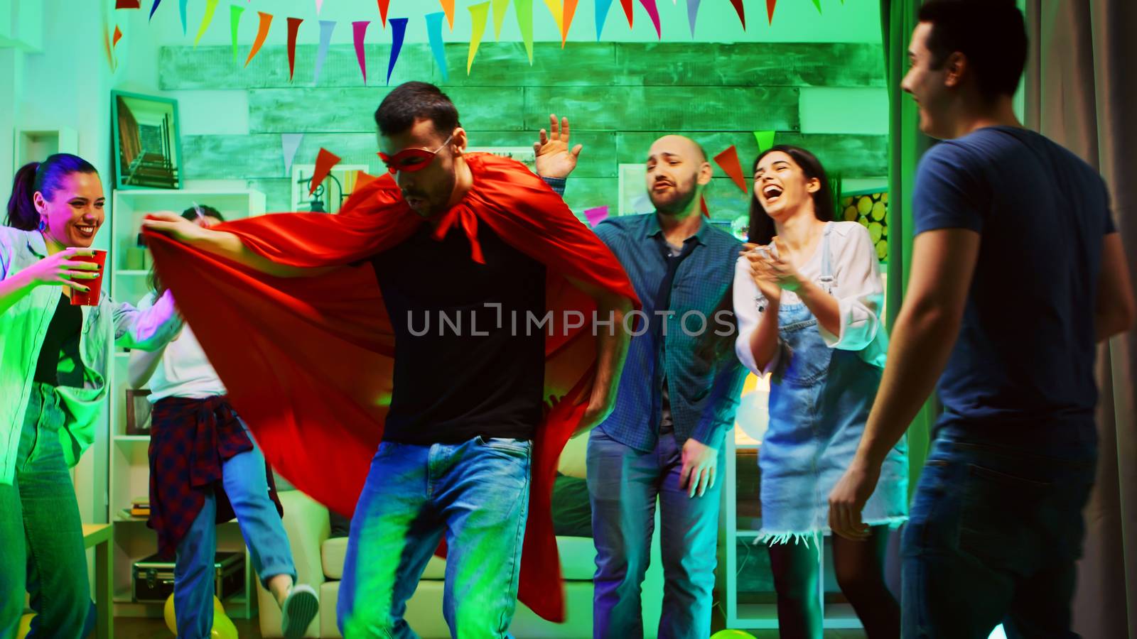 Excited young man wearing a superhero red cape at the party with his friends. Wild college party with neon lights, disco ball and alcohol