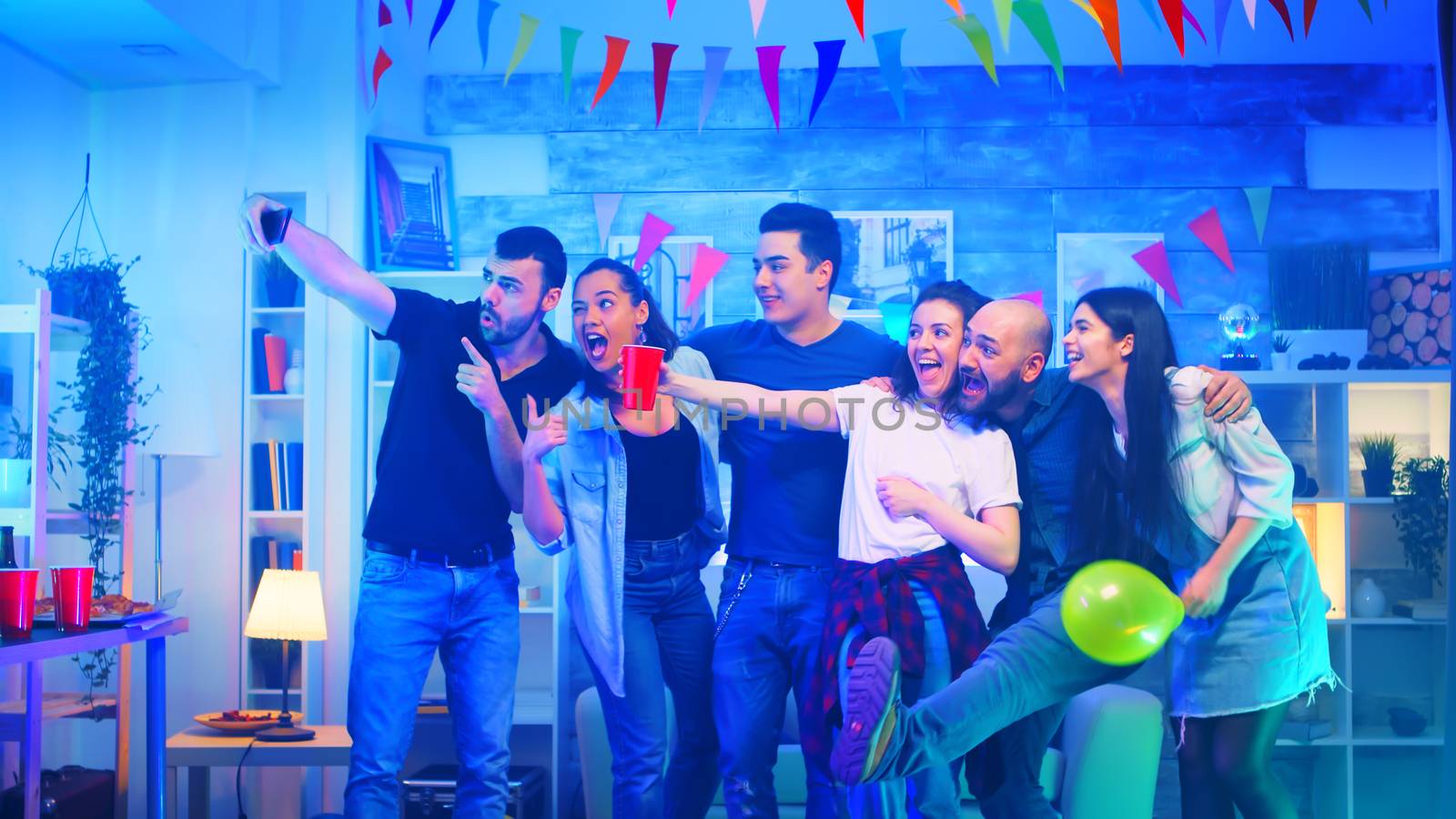 Group of cheerful young people at a party in an apartment with neon light taking a selfie.