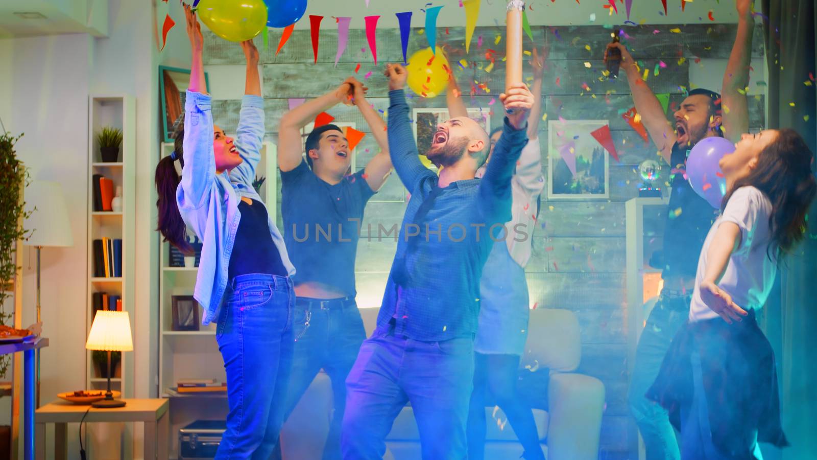Attractive young man throwing confetti while partying with his group of friends in a room with neon lights, disco ball and alcohol. Slow motion shot