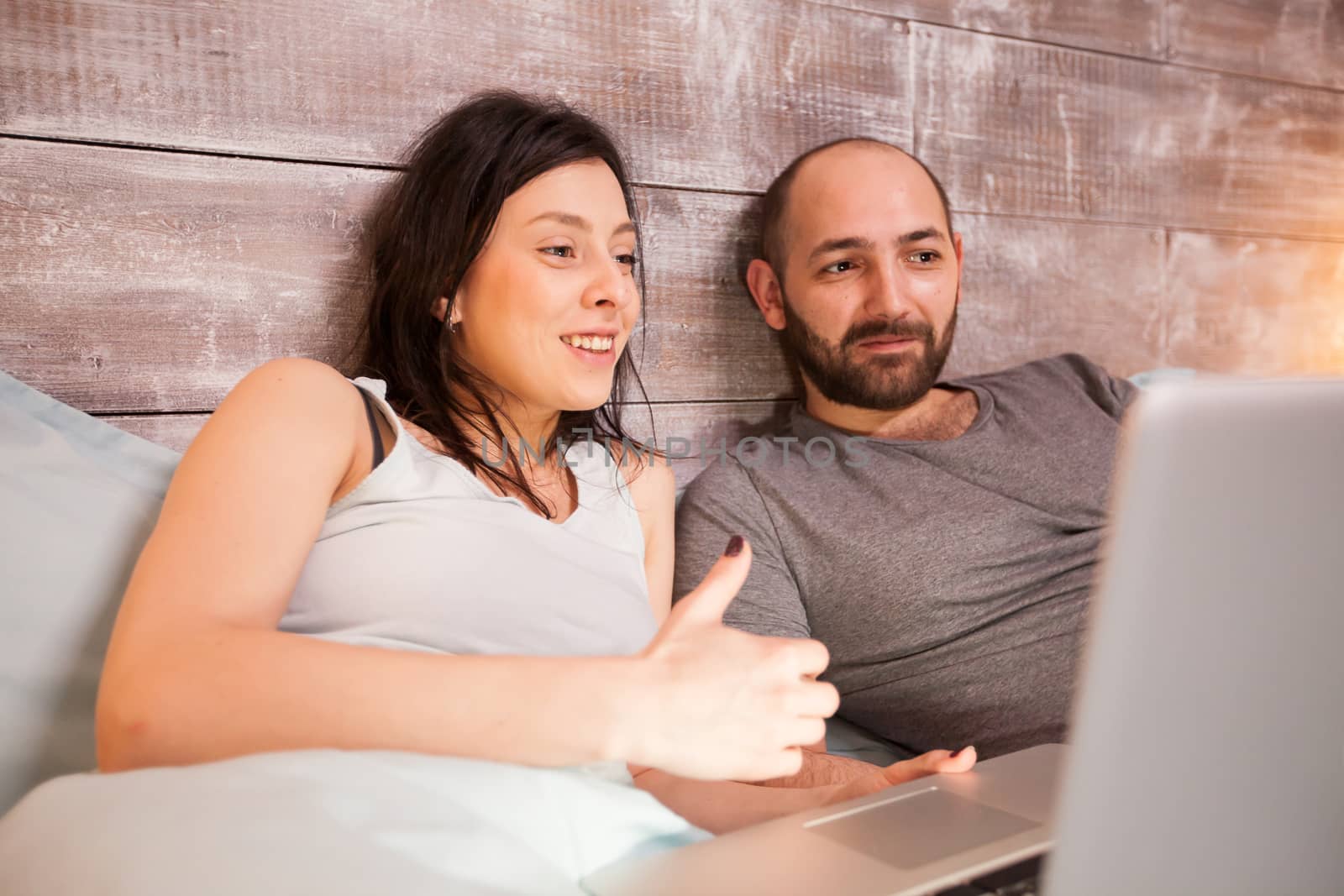 Cheerful young couple in pajamas while watching a movie on laptop laying in bed.