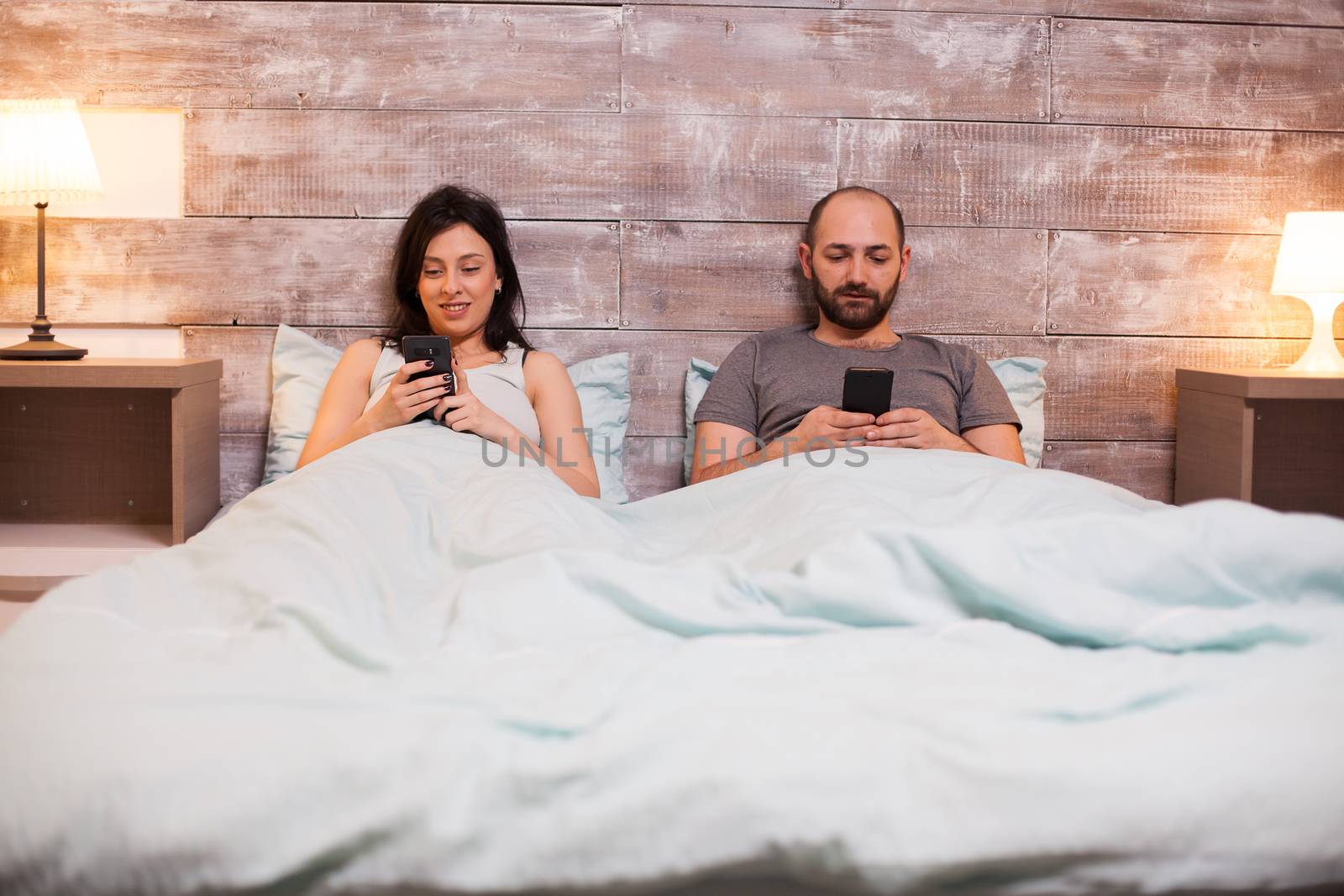 Bored husband browsing on smartphone before bedtime. Beautiful wife in pajamas smiling.