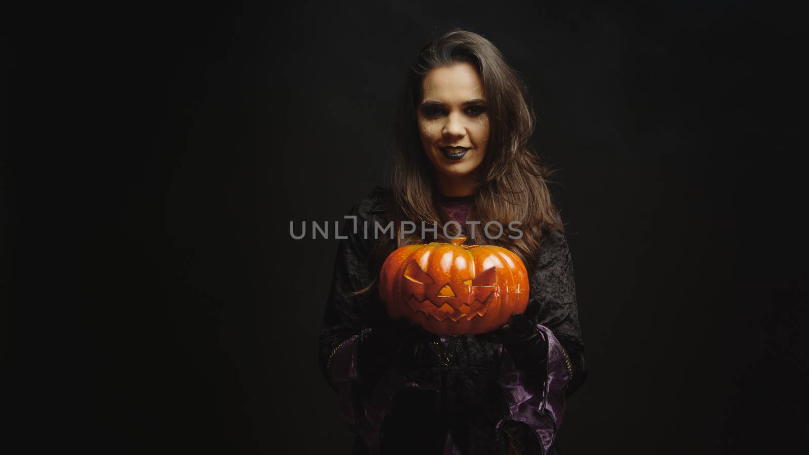 Young woman dressed up like a witch holding a pumpkin for halloween looking at the camera over a black background.