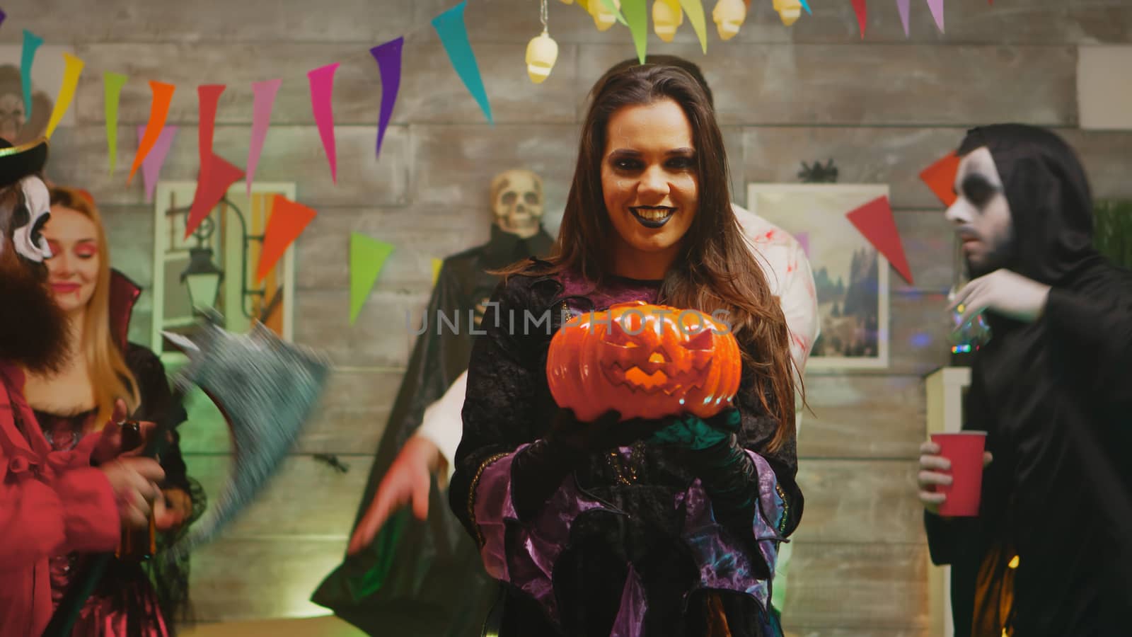 Beautiful young woman dressed up like a scary witch with a pumpkin at a halloween party with her friends.