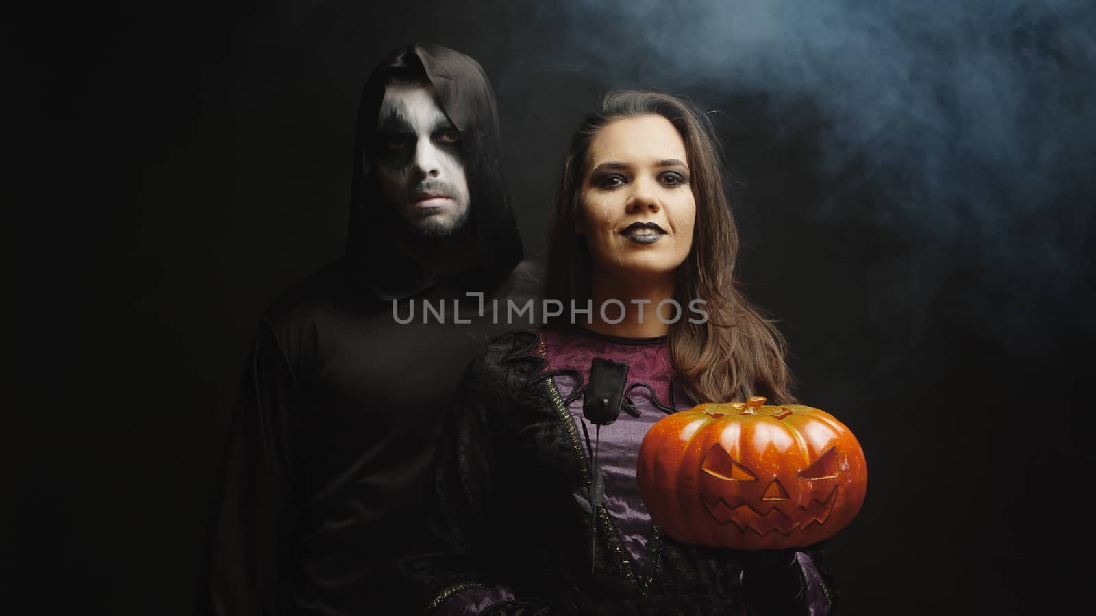 Young woman dressed up like a witch holding a Jack o Lantern for Hallowee next to a dark grim reaper over a black background