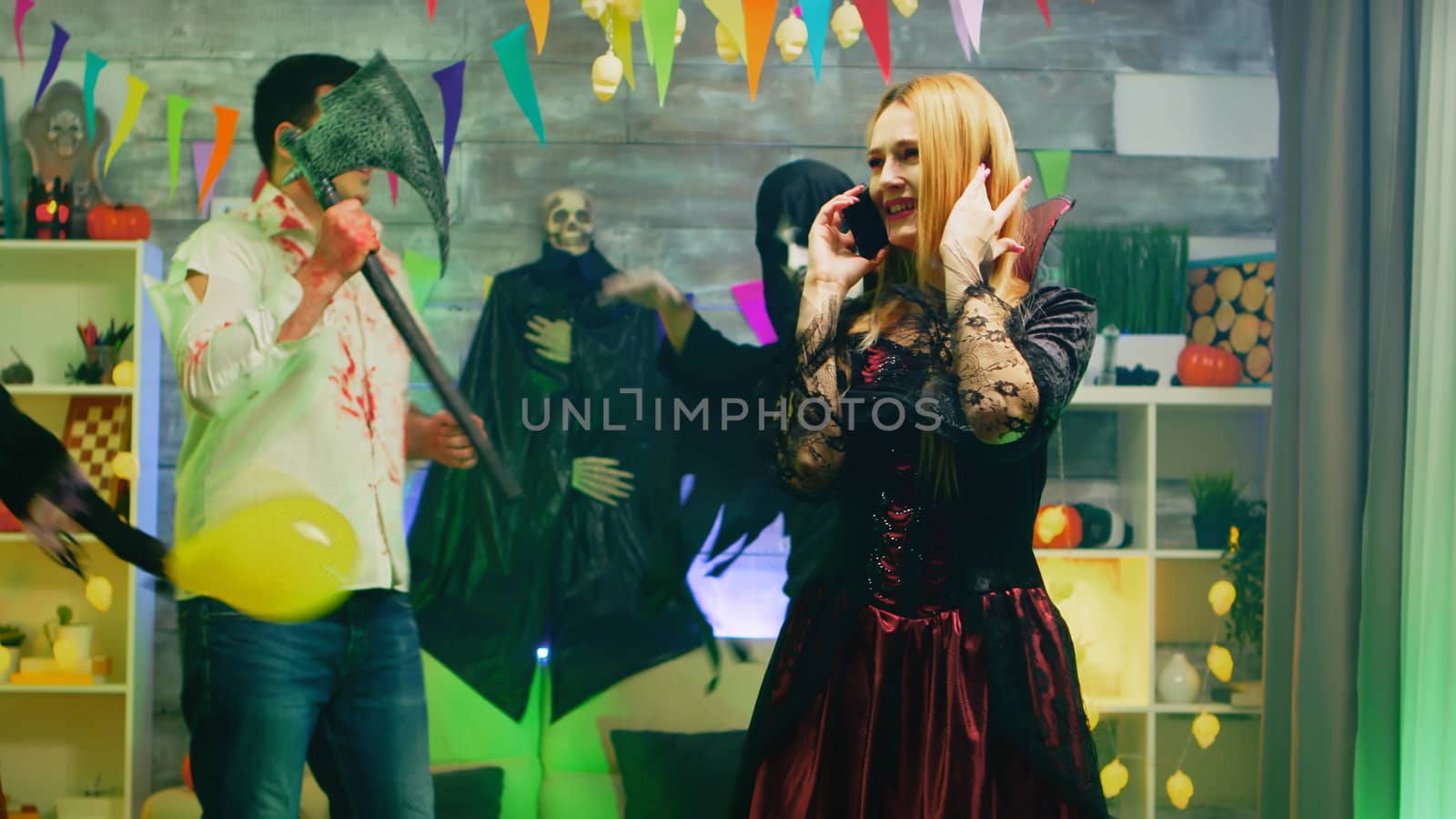 Beautiful woman disguise as a witch talking on her phone at halloween celebration in a room with dancing people