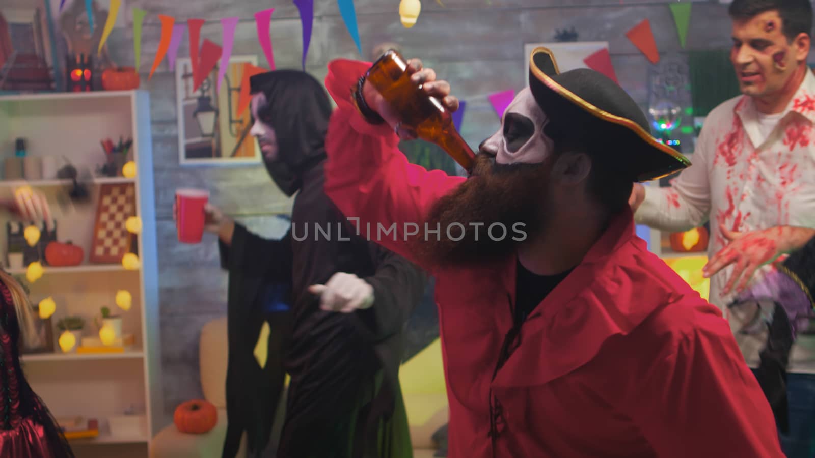 Handsome pirate with an axe drinking beer celebrating halloween with his scary friends dancing in decorated room.