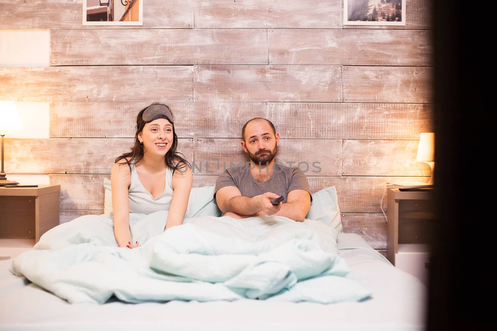 Married caucasian couple in pajamas relaxing watching tv in comfortable bed.