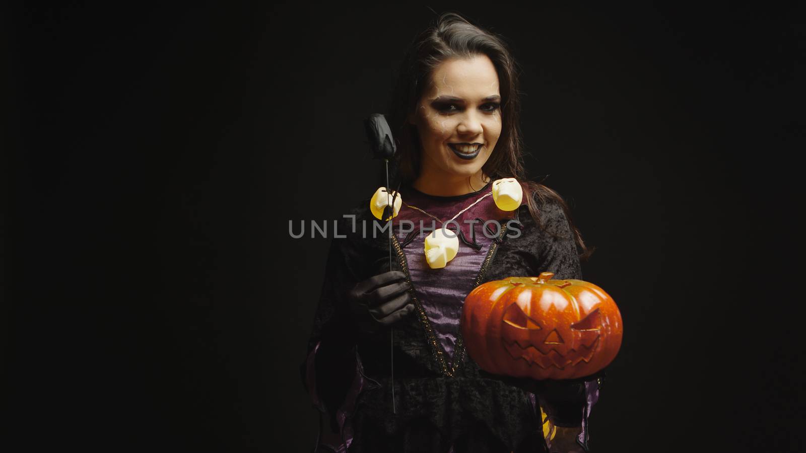Smiling witch dressed up for halloween holding pumpkin over black background.