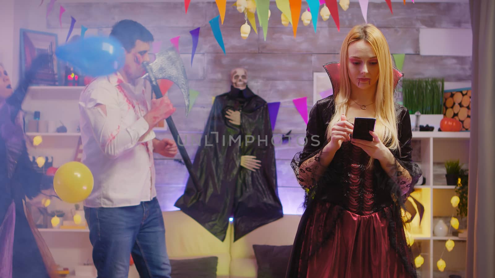Beautiful woman dressed up like an enchantress using her smartphone at halloween party with people dancing in the background.