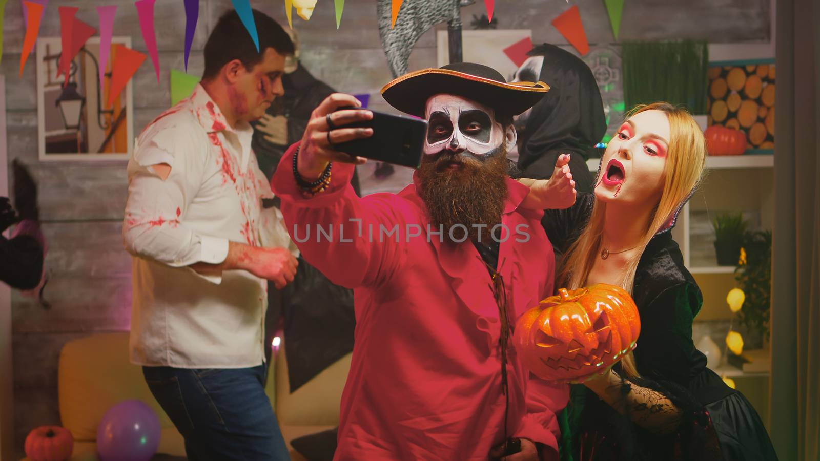 Spooky bearded pirate taking a selfie with beautiful repear woman at halloween party while other are dancing in a decorated house