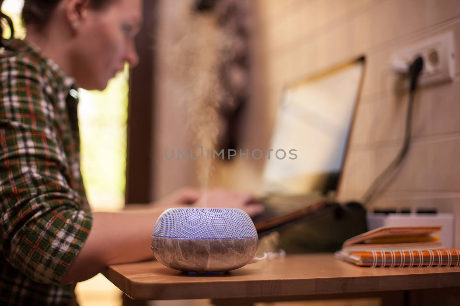 Essential oil diffuser with purple led vaping while woman working on laptop. Covid-19 quarantine.